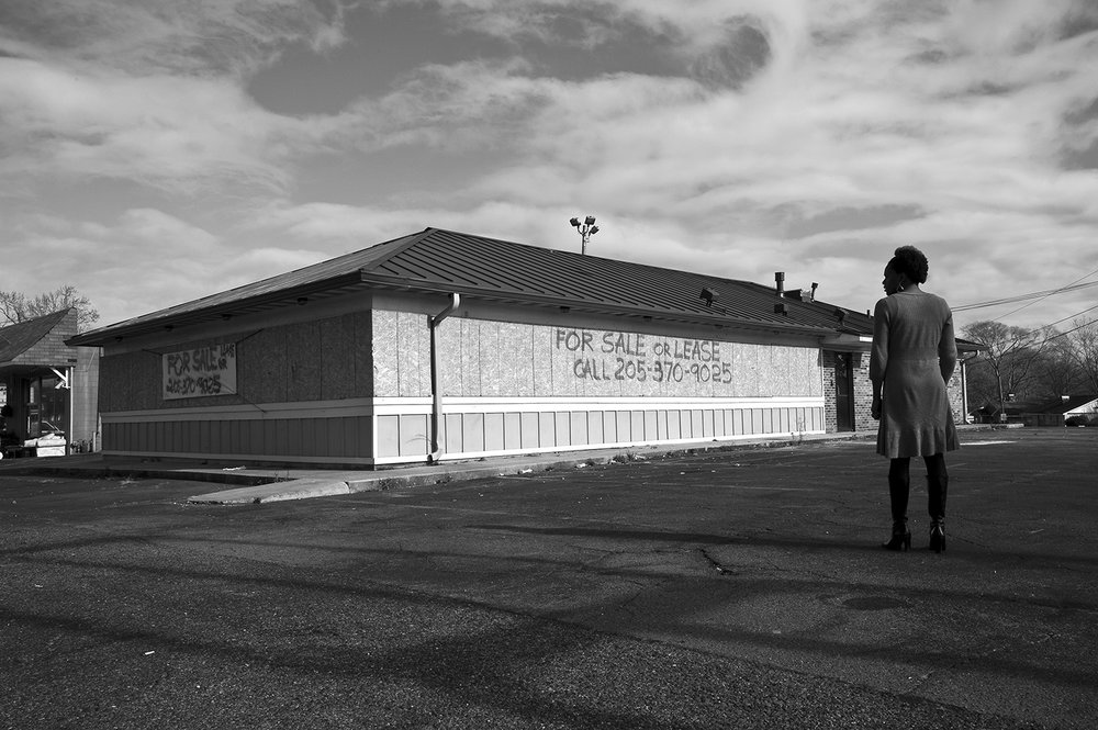 Black and white Image. Woman stands facing an abandoned Pizza Hut. The windows are boarded and state "For sale or lease" in spray paint.