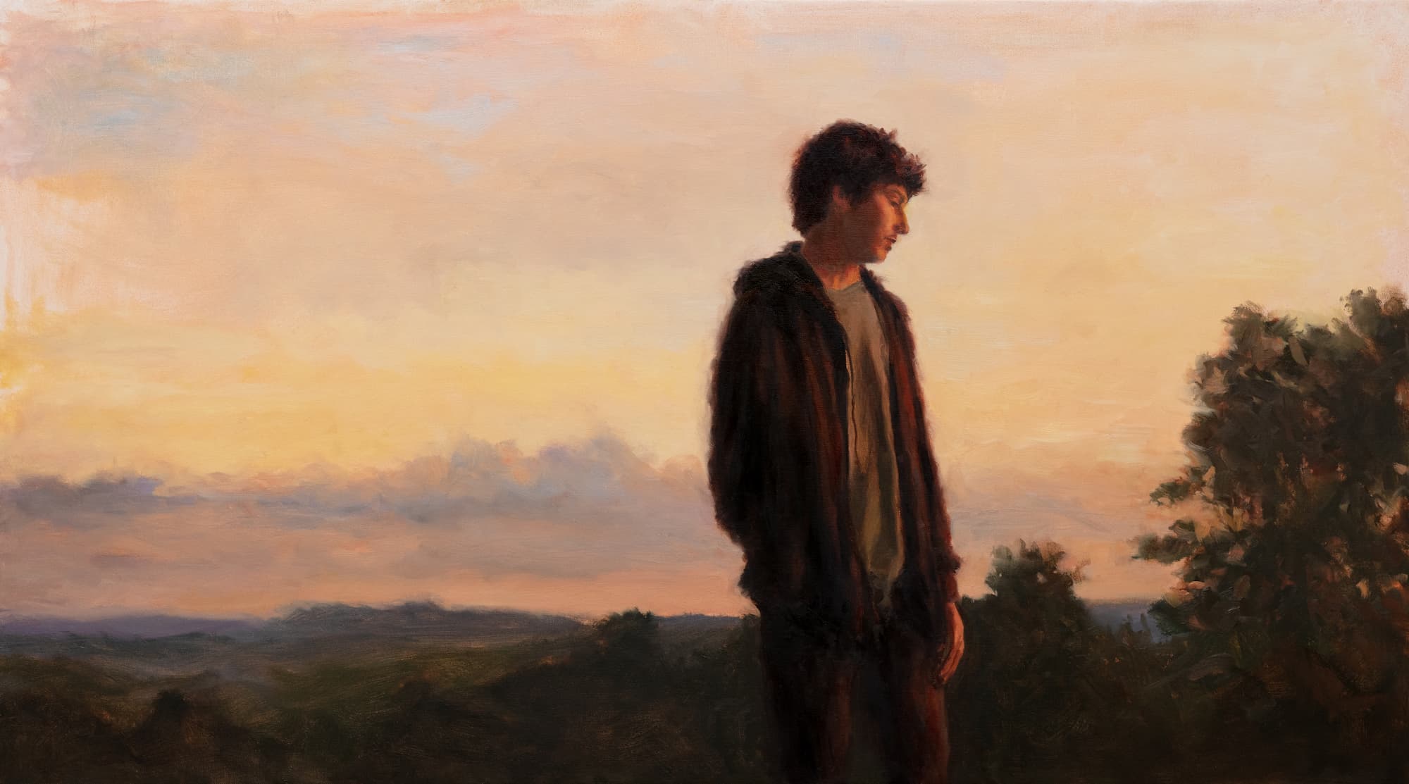A boy stands in a field with a beautiful sunset behind him. 