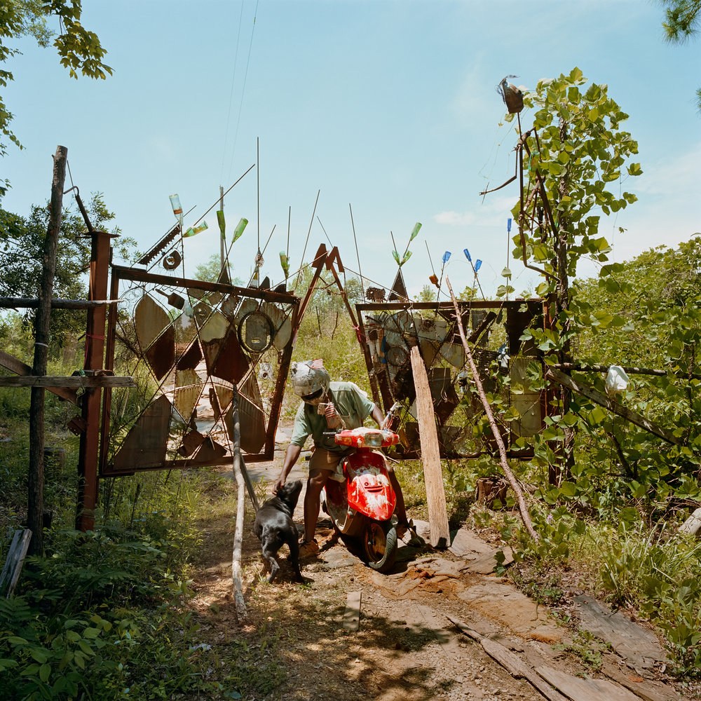 an leans from his motorbike to pet his dog on the head. Behind them is an artistic fence decorated with bottles and scrap metal. 