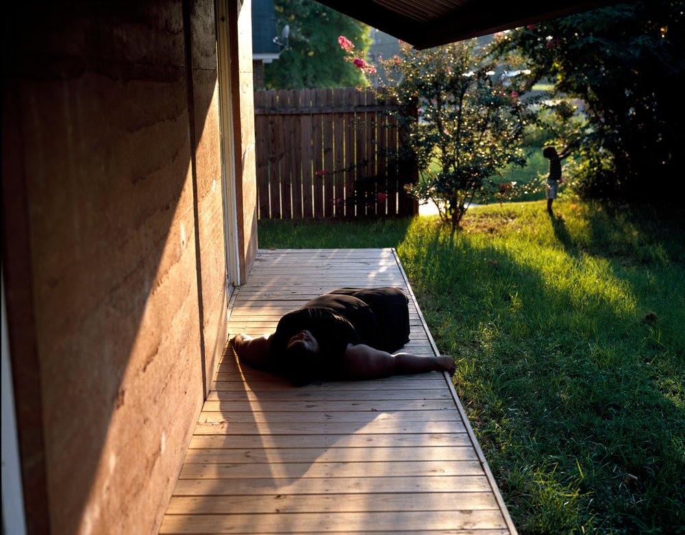 Woman lays, back down, in sunbeam on porch. In the background a small child reaches to grab a treebranch.