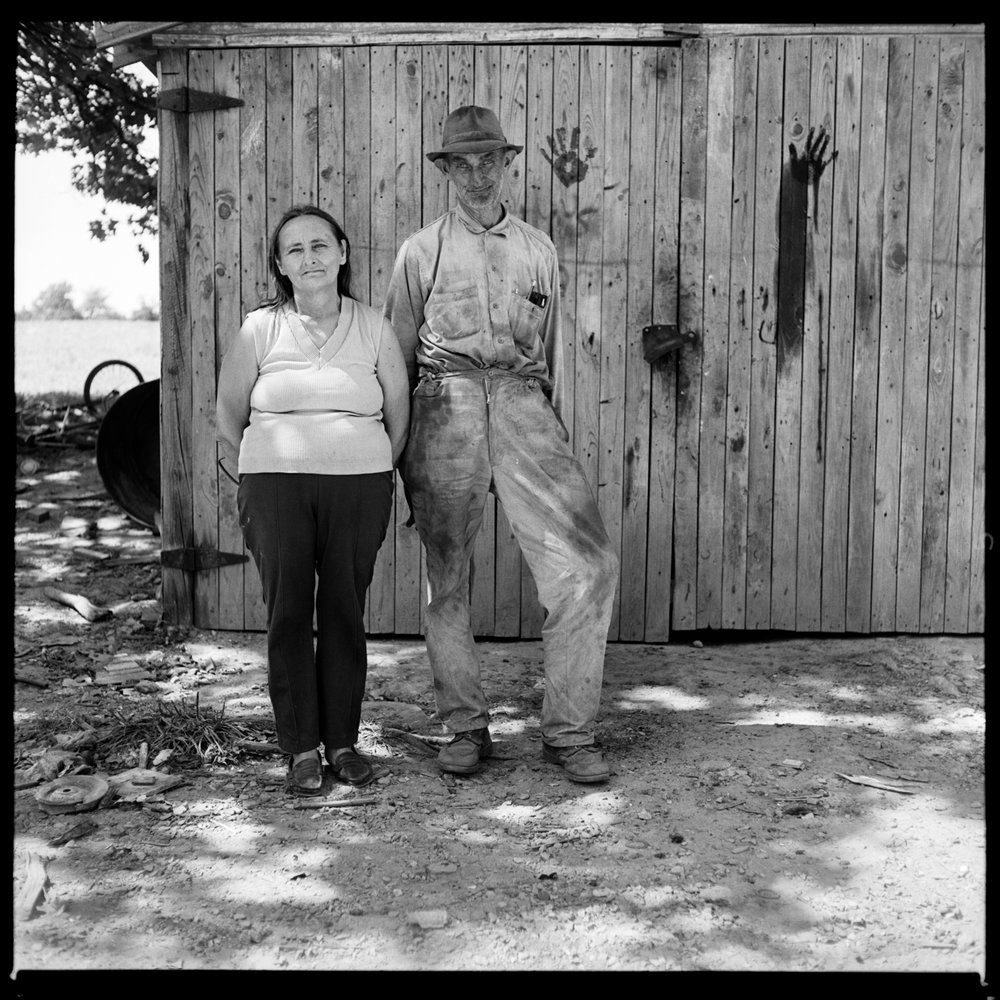 Black and white image of man and woman that stand with their hands behind their backs. Behind them is a wooden shed that has human handprints on either side of the door. 