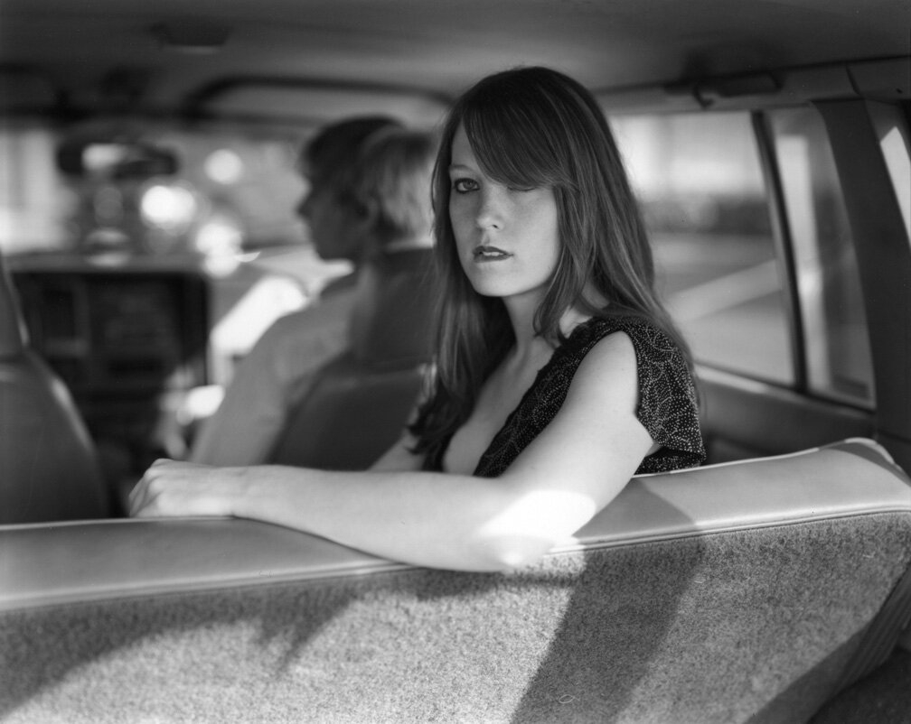 Black and white image of woman in a car. She looks over her shoulder into the camera, slightly biting her lip. 