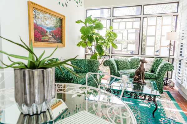 A view inside the sun room reveals green floral love seats, green floral accent rug and a clear round rod iron table.