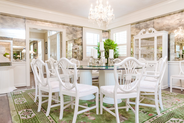 A glass-top, round dining table is surrounded by white-floral cushioned chairs set underneath a hanging crystal chandelier and accented by a white china cabinet with crystal.