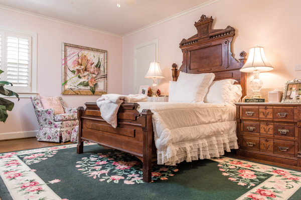 A centered cherry wood bed, along with cherry nightstands adorn the wall when you peer in to the pink-painted bedroom decorated with a green and pink floral rug and floral chair.