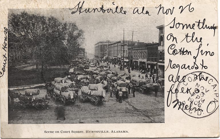 Black and white print showing multiple wagons loaded with cotton in the court square in Huntsville, Alabama. Postmarked November 03, 1906.