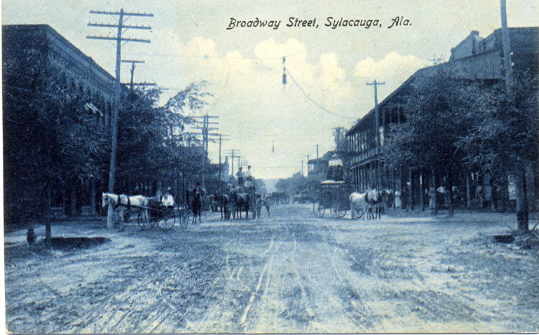 Black and white print of unpaved Broadway Street in Sylacauga, Alabama showing two-story commercial buildings. Postmarked on February 19, 1909.