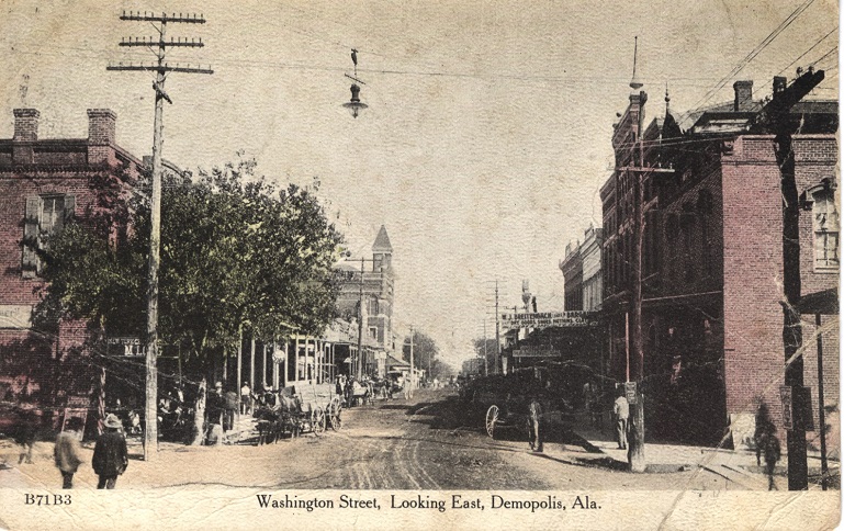 Mostly black and white print of unpaved Washington Street in Demopolis, Alabama showing one and  two-story commercial buildings. Postmarked October 11.