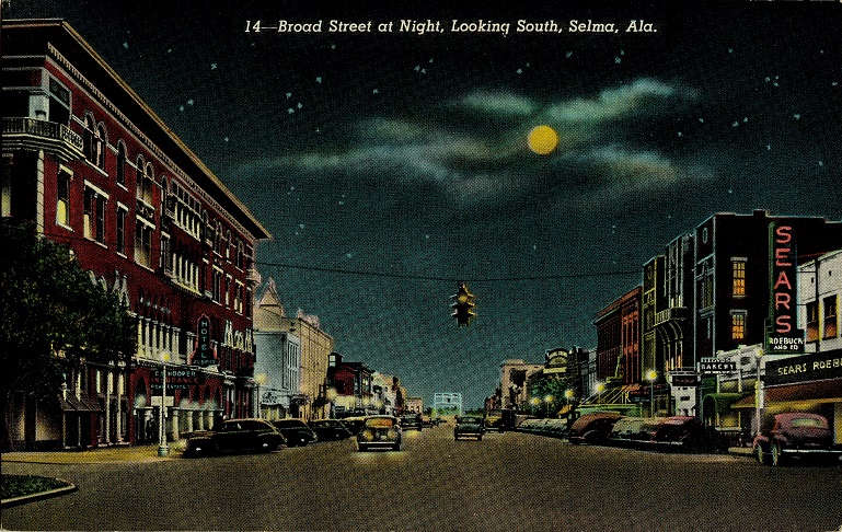 Color print of the Edmund Pettus Bridge and multi-story commercial buildings on Broad Street in Selma, Alabama at night.